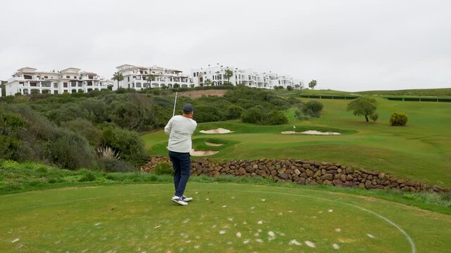 Golfer in white sweater prepares to swing on lush green golf course with luxury villas and sand traps in the background