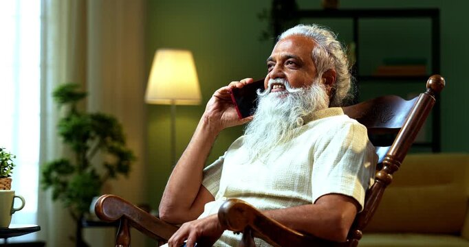 Indian mid age bearded man attending voice call using smartphone sitting on relaxing chair at home, stylish mature male speaking on mobile phone in modern interior lifestyle setting