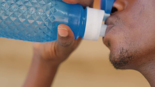 Black man runner sipping from blue sport bottle, closeup on lips and jaw, firm fingertip grip on nozzle, droplets on skin, urgent hydration during steady pace, gritty texture emphasizes effort