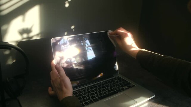 Man hands wiping dust off laptop computer screen with wet wipe while sitting at desk in dark room with sunlight, performing regular electronic device maintenance and care. Male cleaning laptop screen