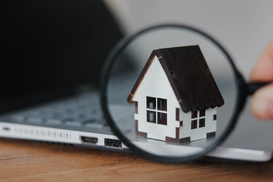 Miniature house viewed through magnifying glass on laptop. Concept of real estate search, property inspection, online housing, mortgage analysis and digital realty services.