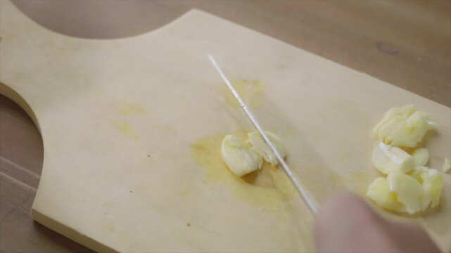 Close-up view of woman's hands with a knife crush the garlic on a chopping board, preparing ingredients for cooking. food preparation and healthy eating concept