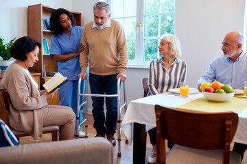 Obraz premium Middle aged Black woman caregiver assisting senior Caucasian man with disability using walker while two senior Caucasian women and senior Caucasian man sitting at table observing interaction