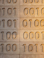 Zeros and ones of binary code are carved as a relief pattern into natural stone cladding, representing low-level programming languages and the foundation of computer science.
