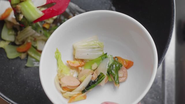 Closeup shot of hands pouring freshly cooked chinese cuisine capcay or chop suey in a white ceramic bowl with a spatula