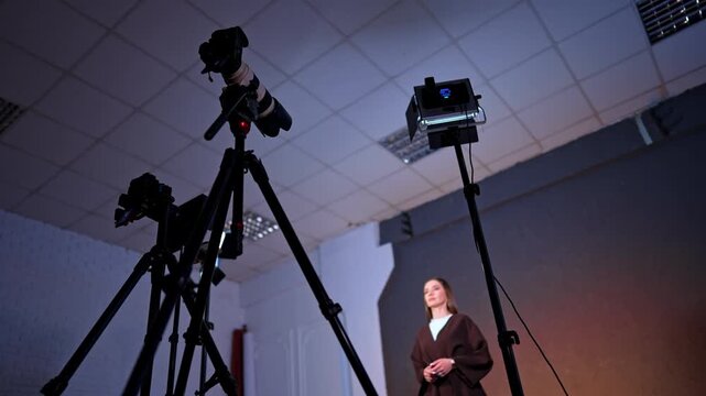 Cameras on tripods and soffits are directed on the woman talking and gesturing. Low angle view. Studio backstage.