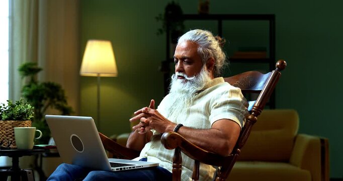 Indian mid age bearded man working on laptop sitting on relaxing chair at home, stylish mature male typing on computer in modern interior setting with wireless digital technology lifestyle