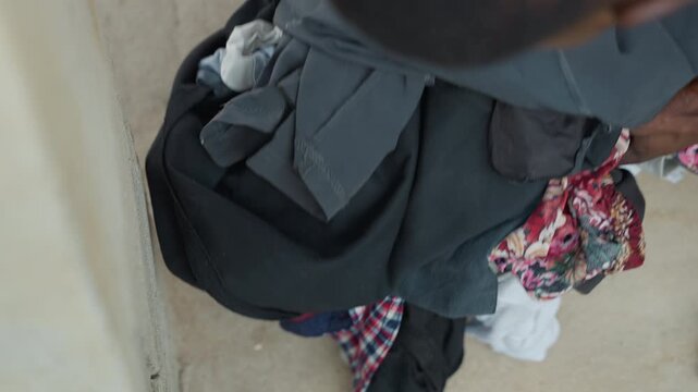Black laundry pile on floor, child folding clothes near concrete steps inside house, hands sorting towels and shirts, muted tones and textured fabric, candid overhead angle conveying quiet domestic