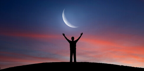 Silhouette of a man with raised hands standing on a hill under a crescent moon at sunset. Elements of this image furnished by NASA.