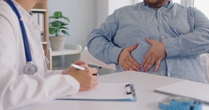 Doctor listens as a patient with obesity shows pain in the abdomen during a clinic consultation. Obesity patient reports stomach pain to physician sitting at desk in medical office.