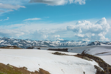 Scenic alpine landscape with sunlit snowfield with snow cornice above abyss against large snow-capped mountain range under cloudy sky. Snowy field near rocky precipice edge. High mountains in sunlight © Daniil