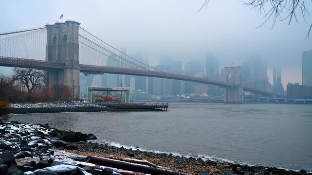 Janes Carousel and Brooklyn Bridge in misty winter weather. View of glass enclosed carousel near Brooklyn Bridge with snowy shoreline and foggy city background at Dumbo.