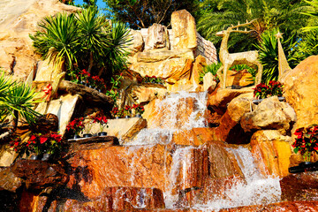 Waterfall flows down rocks surrounded by plants in sunny outdoor area in garden setting. Water cascades over stones in garden with green plants around in Sunset Town, Phu Quoc Island in Vietnam.  © MaxSafaniuk