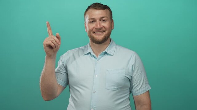 Man points index finger upward in teal studio, showing bare hand and relaxed smile while looking up toward corner; idea optimism.