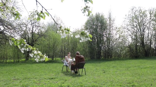 A young couple drinking coffee and having breakfast at a table in the backyard in spring among flowering trees