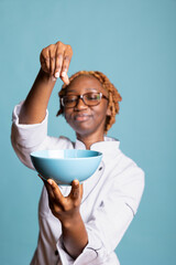 Happy woman chef seasoning cooked food in bowl, finishing dish with care. Professional cook enjoying creative process, preparing flavorful meals with fresh ingredients and condiments.