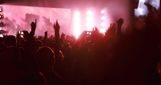 Silhouetted fans raise their arms and hold up mobile phones during a high-energy live performance. Intense red stage lights fill the background.