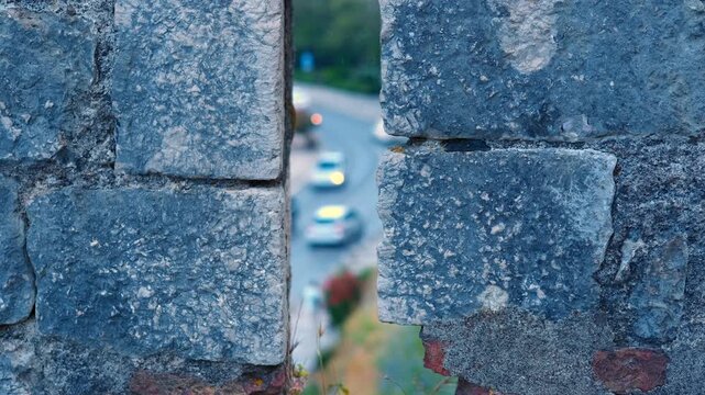 Cars driving on road viewed through castle embrasure. Distant city traffic with cars driving on a winding road, viewed from a high angle perspective through a narrow embrasure in an ancient