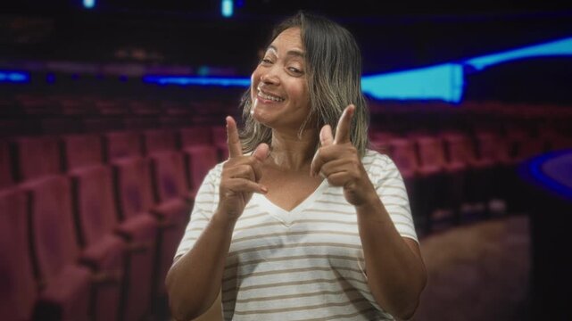 Hispanic woman pointing both index fingers at camera with smiling expression amid theater seats in studio; playful confidence.