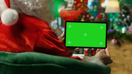 Fototapeta premium Santa Claus reads letters from kids on isolated screen device in Christmas ornaments adorn workshop. Old costumed man in holiday season ambiance holding chroma key tablet, camera A