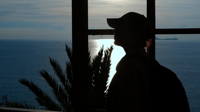 Silhouette of a woman traveler wearing a cap looking at the sunset over the sea. Young thoughtful woman silhouetted against the setting sun watching the calm sea from a window with a palm tree