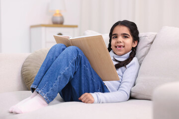 Fototapeta premium Cute little girl reading book on couch at home