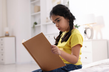 Fototapeta premium Cute little girl reading book on bed at home