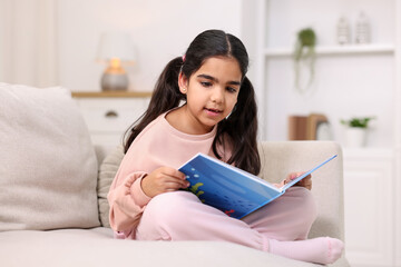 Fototapeta premium Cute little girl reading book on couch at home