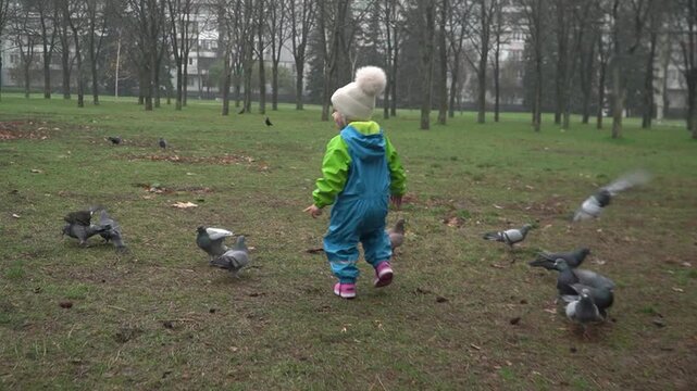 A girl in an autumn jumpsuit runs toward a flock of pigeons to scare them away. She turns to the camera, smiling, satisfied. Slow motion.