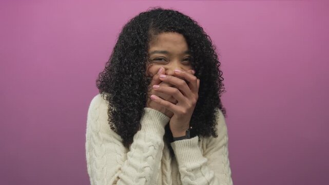 African american woman covers mouth with both hands gesture in vibrant pink studio setting; amusement joy.