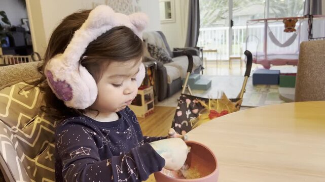 Toddler wearing noise cancelling earmuffs feeding herself during mealtime in real family home environment