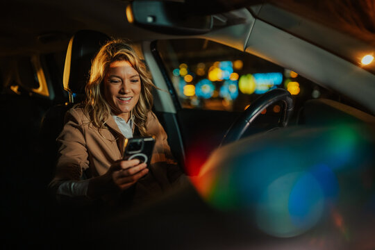 Woman smiling, using smartphone inside a car at night, traveling in a ride share vehicle through city lights