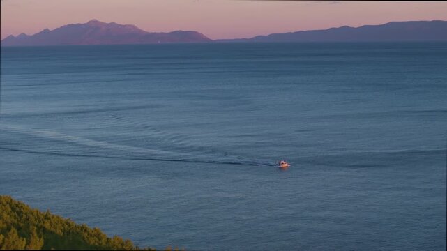AERIAL: A small boat leaves a long trail as it sails across the calm blue Adriatic Sea beneath pink sky. Layered mountains and rugged Dalmatian coastline rise in the distance under soft evening light