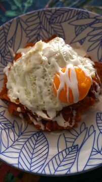 Close-up of red chilaquiles topped with a sunny-side-up fried egg and Mexican crema on a blue leaf pattern bowl.
