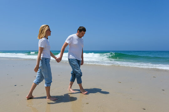 Couple walking barefoot on the beach, hand in hand, with ocean waves and a blueCouple walking barefoot along the beach, hand in hand, with ocean waves and a clear blue sky in the background.