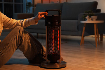 Young woman warming hands near electric heater in living room at evening