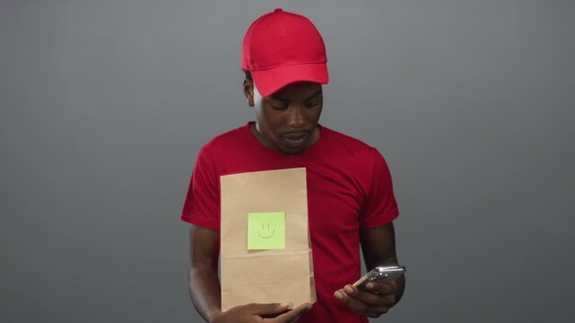Young man holding smartphone and paper bag with smiley note while looking down in gray studio; curiosity.