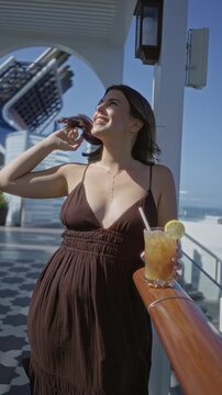 Woman brunette smiles and touches hair while holding iced tea with lemon at cruise terrace railing, visible cleavage and sunlit deck; sunny relaxation.