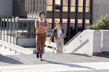 College students walking campus plaza carrying textbooks and folders, wearing beanie and headphones
