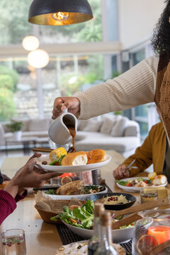 Adults passing dishes, pouring gravy from white pitcher onto plate with rolls at wooden table