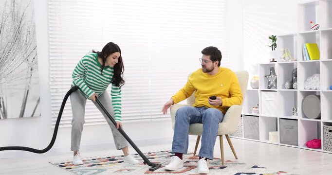 Gender inequality concept. Man with smartphone resting in armchair while his wife doing household chores indoors