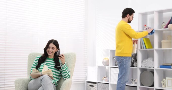 Gender inequality concept. Woman with glass of wine reading book while her husband doing household chores indoors