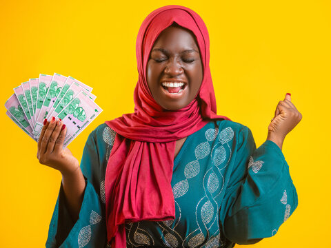 A joyful Muslim woman in a red hijab and teal dress celebrates while holding a fanned stack of green 100 Zimbabwe Dollar banknotes against a bright yellow background, symbolizing financial success.