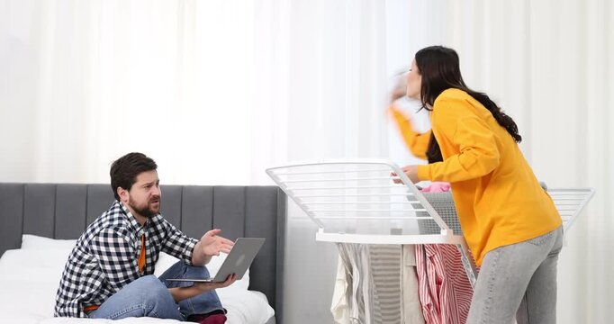 Gender inequality concept. Woman hanging clean laundry on drying rack while her husband using laptop indoors