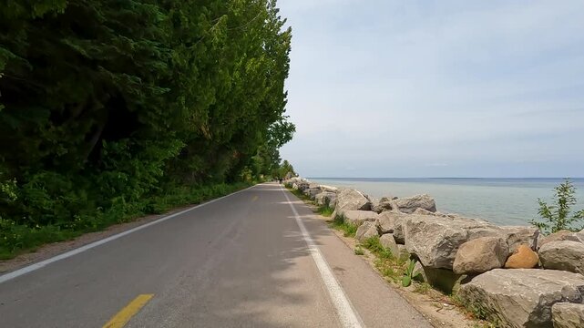Slow-motion POV biking M-185 on Mackinac Island, MI. The paved coastal road stretches ahead with cyclists and pedestrians, flanked by a rocky Lake Huron shore and green forest.