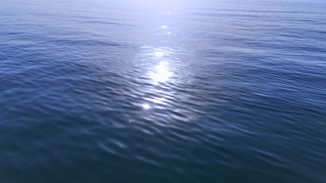 Cinematic drone flight over calm ocean water with bright sun glare and soft rolling ripples, minimal blue marine landscape background in serene daylight