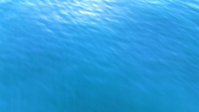 Top view drone footage gliding above deep blue sea with sparkling sun reflection and soft wave patterns, peaceful ocean background in bright natural light