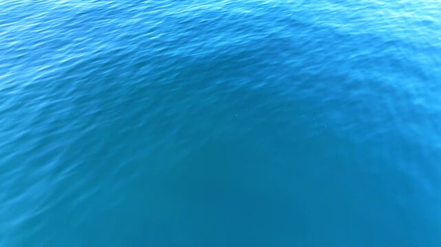 Top view drone footage gliding above deep blue sea with sparkling sun reflection and soft wave patterns, peaceful ocean background in bright natural light