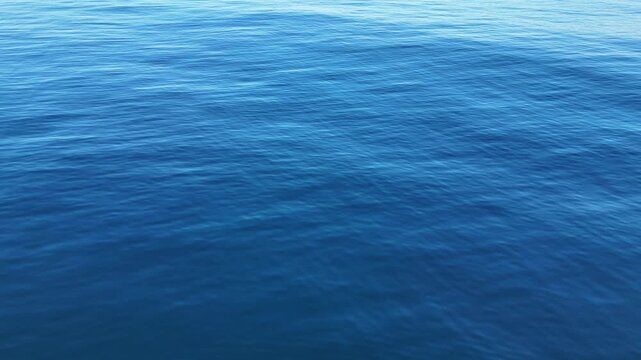 Aerial drone shot flying over calm blue ocean surface with shimmering sunlight reflection and gentle ripples, cinematic seascape background in natural daylight