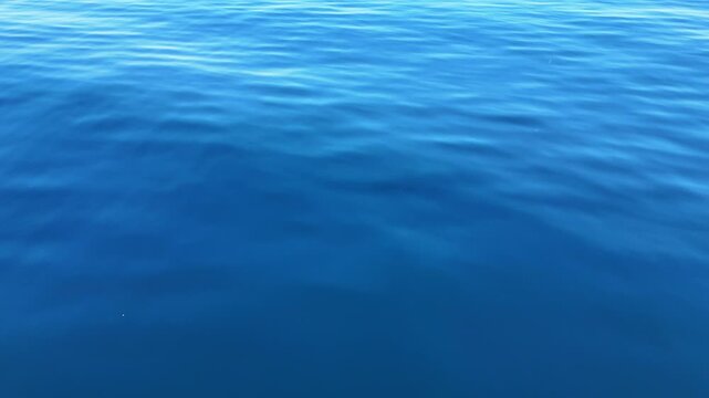 Aerial drone shot flying over calm blue ocean surface with shimmering sunlight reflection and gentle ripples, cinematic seascape background in natural daylight
